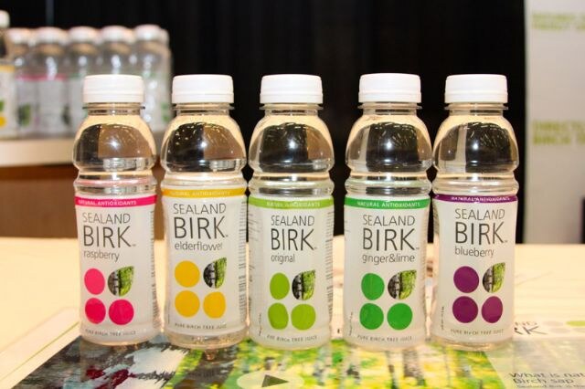 Five clear bottles of Sealand Birk birch tree juice in raspberry, elderflower, original, ginger and lime, and blueberry stand in a row on a table.