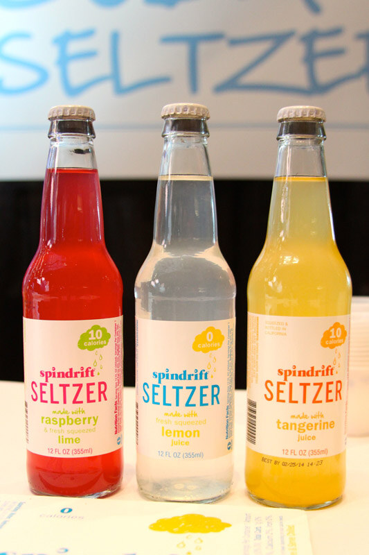 Three glass bottles of Spindrift Seltzer in raspberry lime, lemon and tangerine flavors stand in a row on a table in front of a banner reading Seltzer.