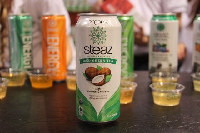 A can of Steaz organic iced green tea with coconut water in front of other Steaz cans and small sample cups on a dark table.