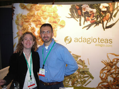 A woman with curly hair and a man in a blue shirt, both wearing green lanyards, stand in front of an Adagio Teas backdrop showing loose tea leaves and dried fruit.