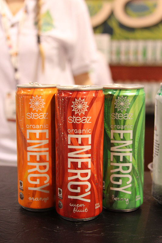 Three cans of Steaz Organic Energy in orange, super fruit and berry stand on a dark counter, with a person in a white shirt standing blurred behind them.