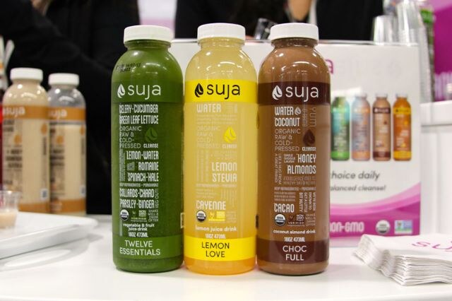 Three Suja bottles named Twelve Essentials, Lemon Love and Choc Full stand on a white counter, with more bottles and a display behind them.