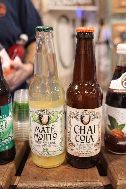 Two glass bottles of Taylor's Tonics, Maté Mojito and Chai Cola, on a wooden crate, with a stack of plastic sample cups and other bottles nearby.