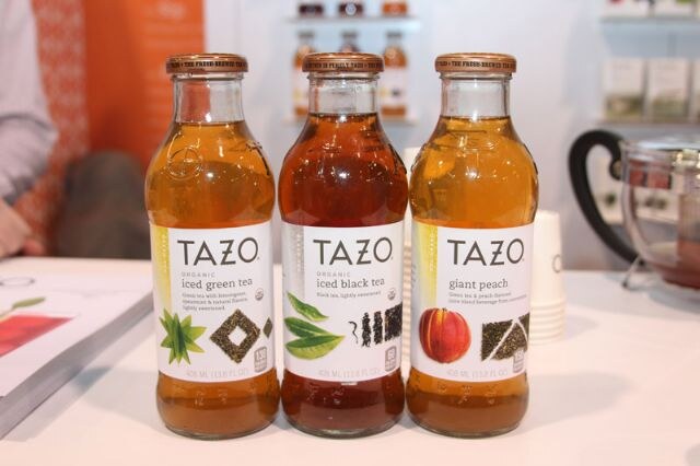 Three glass bottles of Tazo organic iced tea, iced green tea, iced black tea and giant peach, stand on a white table.