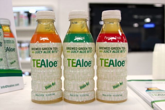 Three TEAloe brewed green tea with aloe bits bottles in Peach, Original and Passion Fruit flavors stand on a counter next to brochures.