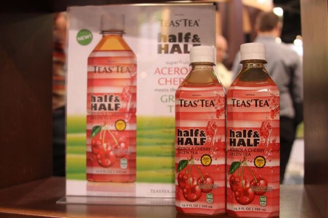 Two bottles of Teas' Tea Half & Half Acerola Cherry Green Tea in front of a sign for the same product on a shelf, with a person blurred behind.