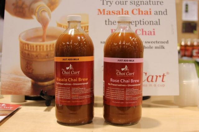 Two bottles of The Chai Cart chai brew, Masala Chai Brew and Rose Chai Brew, stand on a table in front of a sign about masala chai.