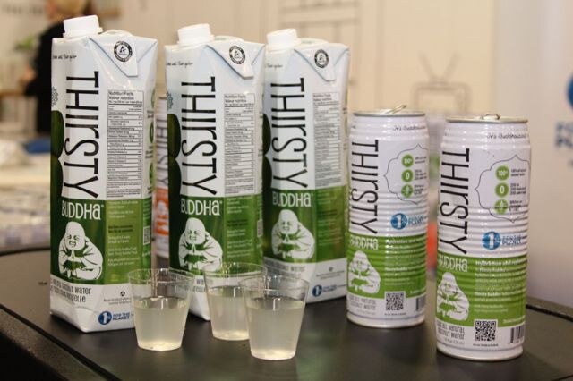 Three Thirsty Buddha cartons and two cans on a dark table, with three small cups of clear drink sample in front.