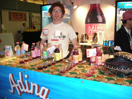 A curly-haired man with glasses stands behind a booth counter with Adina bottles and small cups of red drink samples. A large Adina bottle is displayed behind him.