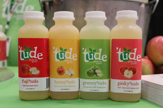 Four bottles of 'tude apple juice in a row on a green table: fuji'tude, honey'tude, granny'tude and pinky'tude, with apples visible at the right.