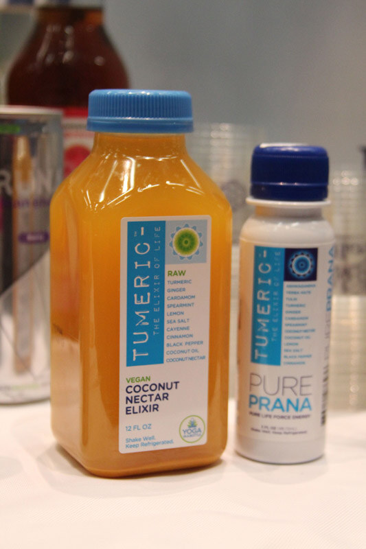 Two bottles on a white table: a Tumeric Vegan Coconut Nectar Elixir in a blue-capped bottle and a smaller Tumeric Pure Prana shot bottle with a dark blue cap.