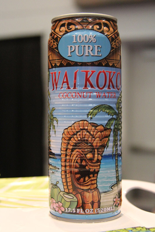 A tall can of Waikoko 100% Pure Coconut Water with a tiki figure, palm trees and beach illustration stands on a white table.