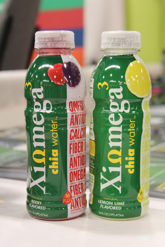 Two green bottles of Xiomega chia water, Berry and Lemon Lime flavors, stand on a white table with printed materials and a colorful backdrop behind them.
