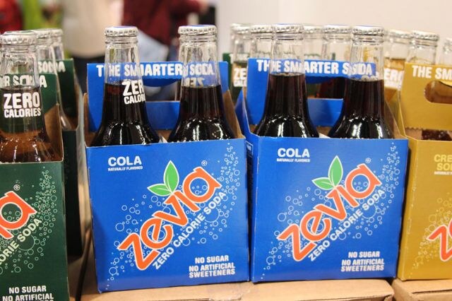 Blue four-pack cartons of Zevia cola zero calorie soda hold dark glass bottles, with green and yellow Zevia packs partly visible on either side.