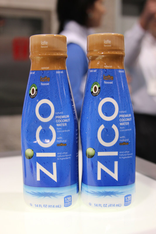Two blue bottles of ZICO latte flavored premium coconut water with tan caps stand side by side on a white surface, with blurred people in the background.