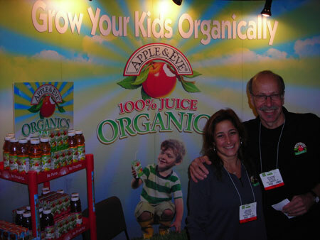 A woman and a man in dark clothing with name badges pose in front of an Apple & Eve backdrop reading Grow Your Kids Organically, with a child photo and juice products on a shelf.