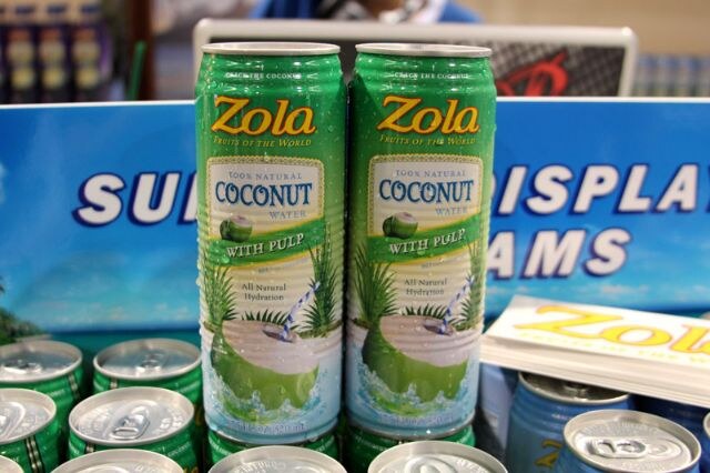 Two tall green cans of Zola coconut water with pulp stand stacked on more Zola cans, in front of a blue display banner with partial text.