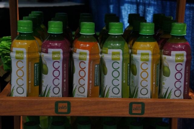 Wooden shelf holding rows of Alo Coco bottles with green caps in mangosteen mango, goji berry lychee, and passion fruit pineapple flavors, with the alo logo on the shelf edge.