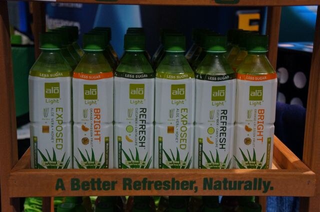 Wooden shelf holding rows of Alo Light aloe vera drink bottles in Exposed, Bright and Refresh flavors, with the slogan "A Better Refresher, Naturally." on the front.
