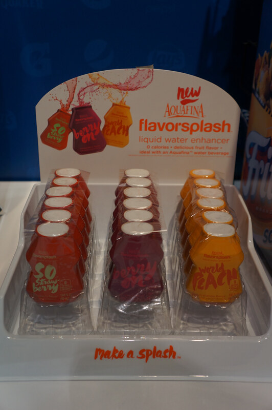 A countertop display of Aquafina FlavorSplash liquid water enhancer in three rows of small bottles in strawberry, berry and peach flavors, with the line Make a splash.