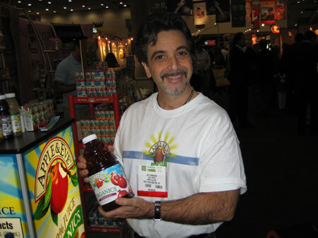 A smiling man with dark hair and a goatee wears a white Apple & Eve t-shirt and name badge. He holds a bottle of juice next to an Apple & Eve display.