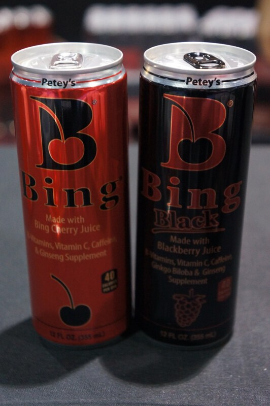 Two 12 fl oz cans labeled Petey's, a red Bing made with cherry juice and a black Bing Black made with blackberry juice, on a gray tablecloth.
