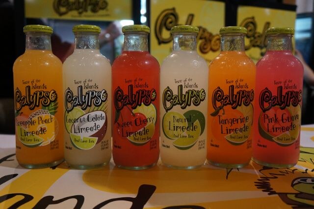 Six Calypso lemonade bottles lined up on a table: Pineapple Peach, Coconut Colada, Sweet Cherry, Natural, Tangerine and Pink Guava Limeade, in front of yellow Calypso signs.