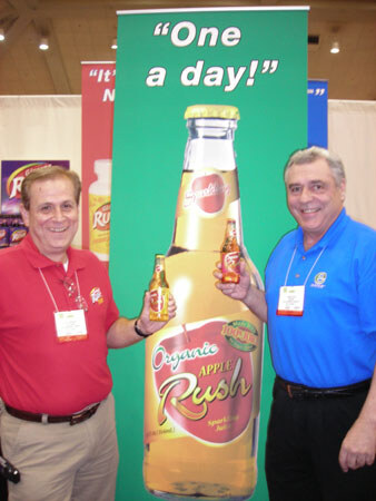 Two smiling men, one in a red shirt and one in a blue shirt, each hold a small Apple Rush bottle beside a tall Organic Apple Rush bottle banner reading 'One a day!'