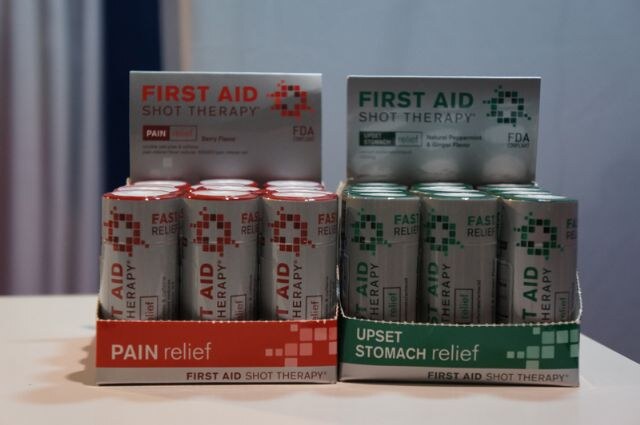 Two display boxes of First Aid Shot Therapy bottles on a table, one labeled Pain Relief in red and one labeled Upset Stomach Relief in green.
