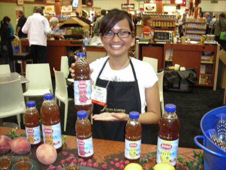 A smiling woman in a black apron with glasses stands behind a table displaying several bottles of Bina iced tea, with peaches, berries and lemons arranged around them.