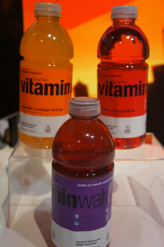 Three Glacéau vitaminwater bottles on clear risers: orange and red labeled bottles in back and a purple labeled bottle in front, against an orange backdrop.