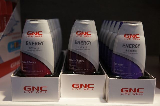 Three boxes of GNC Live Well Energy B Vitamin liquid water enhancer bottles in mixed berry, black cherry and grape flavors, arranged on a shelf.