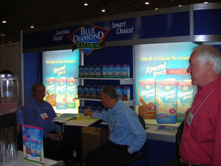 Three men at a Blue Diamond Natural booth with lit Almond Breeze cartons on display. Two are seated, one writes at a table, and a man in a red shirt stands at right.