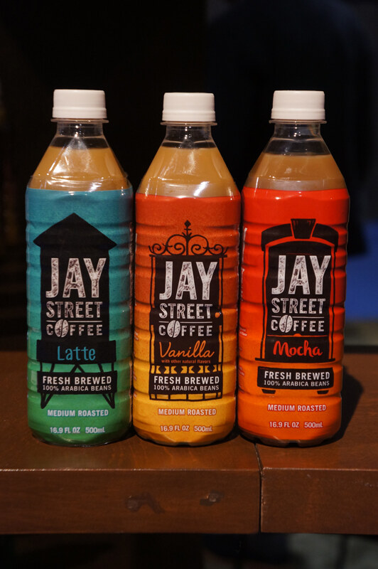 Three bottles of Jay Street Coffee with white caps, in Latte with a green label, Vanilla with an orange-yellow label and Mocha with a red label, on a wooden ledge.
