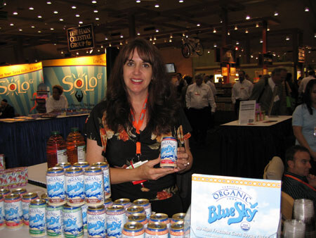 A smiling woman with long dark hair holds a Blue Sky can beside a table of Blue Sky cans, with a Blue Sky sign in front and SoLo banners behind her.