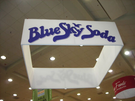 A white square hanging sign reading Blue Sky Soda in blue letters is suspended from a ceiling with recessed lights. A green aisle sign and a red sign are partly visible below.