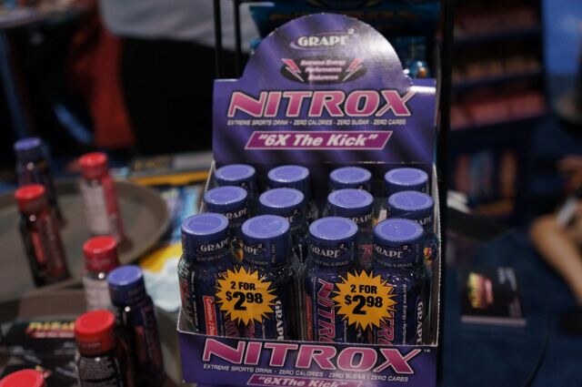 Cardboard display box of purple-capped Nitrox sports drink shots by Grape², with 2 for $2.98 stickers, on a table with other red-capped bottles nearby.