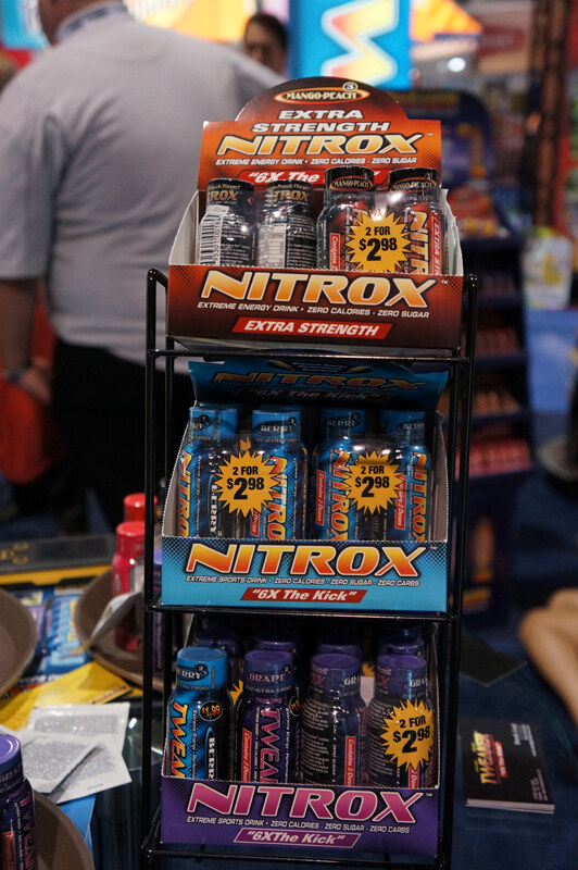 Wire rack holding three boxes of Nitrox shots, Extra Strength on top, blue in the middle and grape at the bottom, with 2 for $2.98 labels; a person stands in the background.