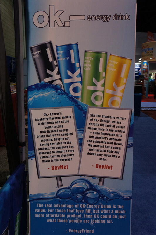 Standing banner for ok.- energy drink showing four colored cans and quotes from BevNet and EnergyFriend about the drink's flavor and value.