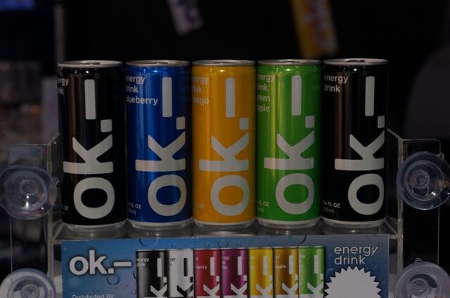 Five slim ok. energy drink cans in black, blue, yellow, green and black stand in a row on a clear acrylic stand above an ok. energy drink sign.