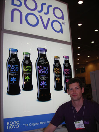A man in a dark purple shirt with a name badge sits below a lit Bossa Nova display showing five bottles of açai juice in different colors.