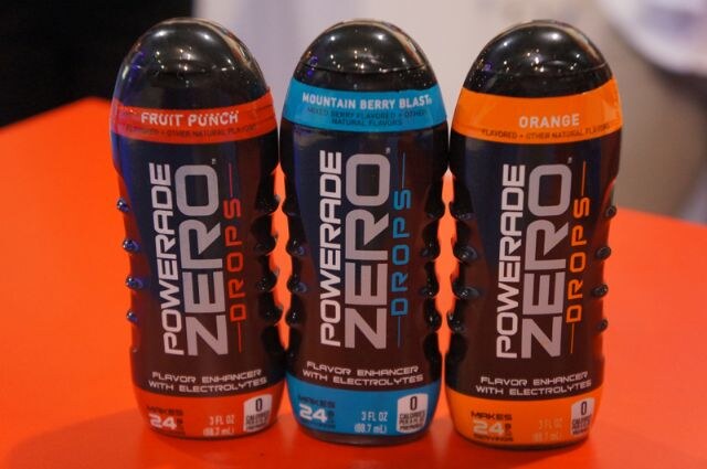 Three Powerade Zero Drops bottles on an orange surface, labeled Fruit Punch, Mountain Berry Blast and Orange, each described as a flavor enhancer with electrolytes.