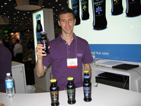 A smiling man in a purple Bossa Nova polo and name badge holds up a dark bottle of açai juice. Three more bottles sit on the counter in front of him, with a lit Bossa Nova display behind.