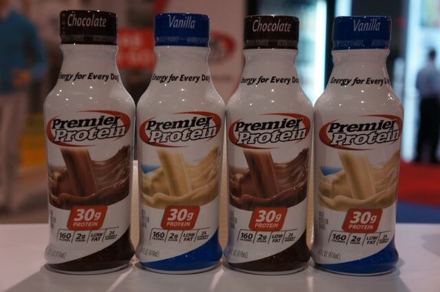 Four Premier Protein shake bottles in a row, alternating chocolate and vanilla, each labeled 30g protein, on a white surface with a blurred show floor behind.