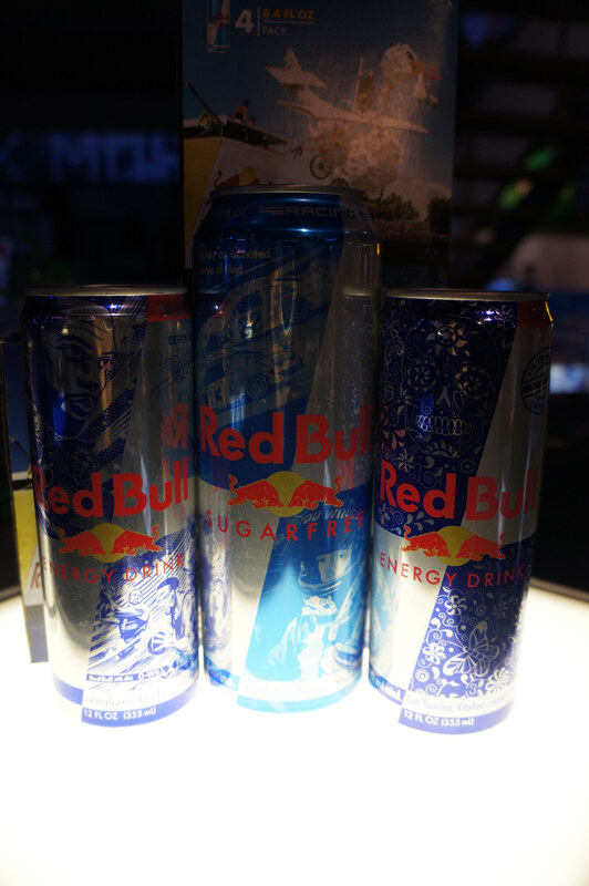 Three Red Bull cans with decorative blue and silver designs, including a Sugarfree can in the center, lit from below in a dark display area.