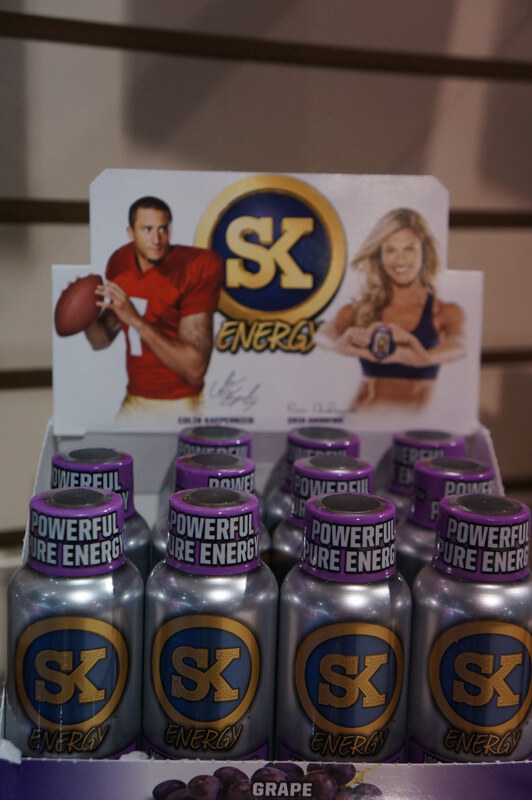 Open display box of SK Energy shots with purple-capped silver bottles reading Powerful Pure Energy, below a header card showing a man holding a football and a woman holding a bottle.