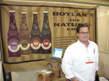 A man in a white shirt and glasses stands in front of a Boylan's banner showing four soda bottles, with a laptop and cartons of product on a table beside him.
