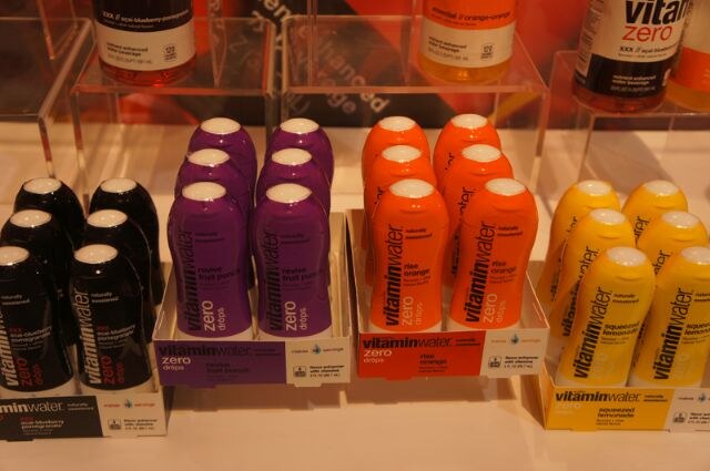 Shelf display of vitaminwater zero drops bottles in black, purple, orange and yellow, grouped in trays by flavor, with larger vitaminwater zero bottles on shelves above.