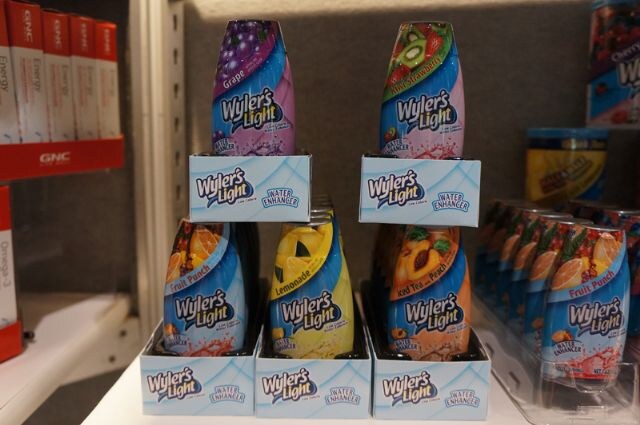 Stacked boxes of Wyler's Light Water Enhancer bottles in grape, strawberry, fruit punch, lemonade and iced tea peach flavors, with GNC boxes on a shelf at left.