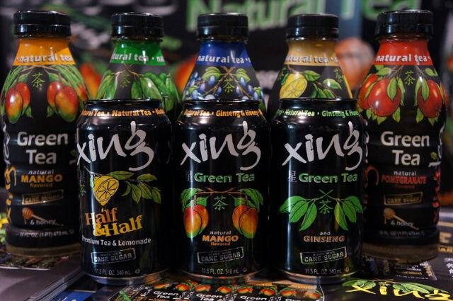 Black Xing green tea cans in half and half, mango and ginseng varieties stand in front of taller natural tea bottles in mango and pomegranate flavors.